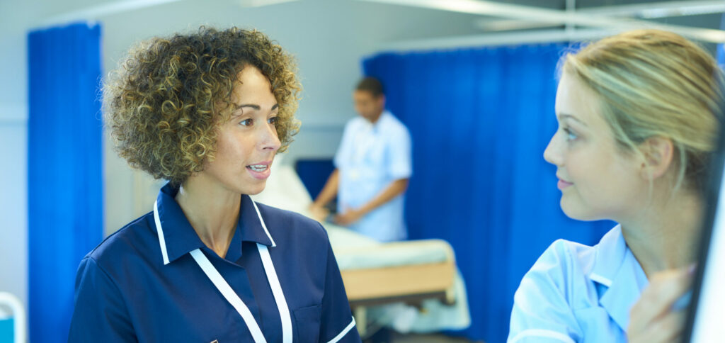 a female ward nurse sister is chatting to a young nurse who is drawing on the wards occupancy chart on a busy hospital ward . In the background beds and cubicles can be seen and a male nurse is attending to one of the hospital beds . The nurse sister is holding a digital tablet and explains to the nurse the situation on the ward .They are all wearing nurse uniforms consistent with the uk and the NHS .