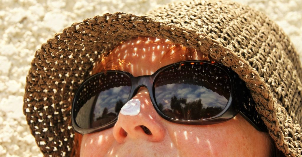 Photograph of a woman wearing sunglasses and a sun hat with a blob of suncream on her nose