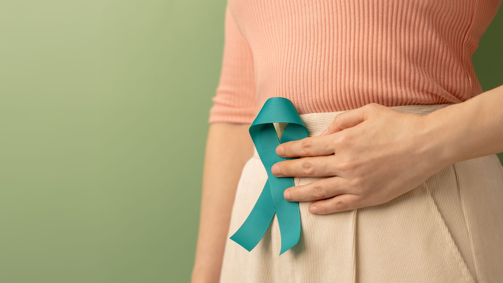 Close-up of a woman wearing a teal awareness ribbon near her abdomen, symbolising ovarian cancer awareness, with a soft green background