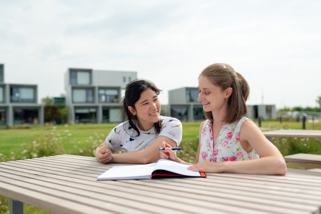 Two young women sitting at a wooden picnic table outdoors, discussing notes in an open book on a sunny day.