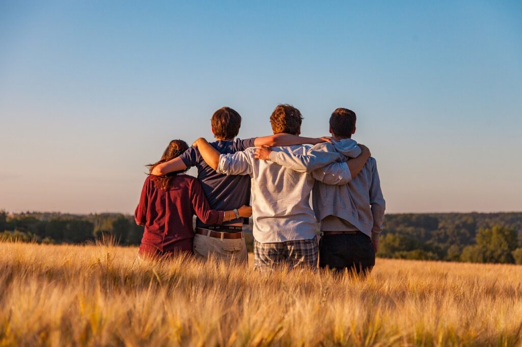 Four friends stand arm in arm in a golden wheat field under a clear blue sky at sunset.