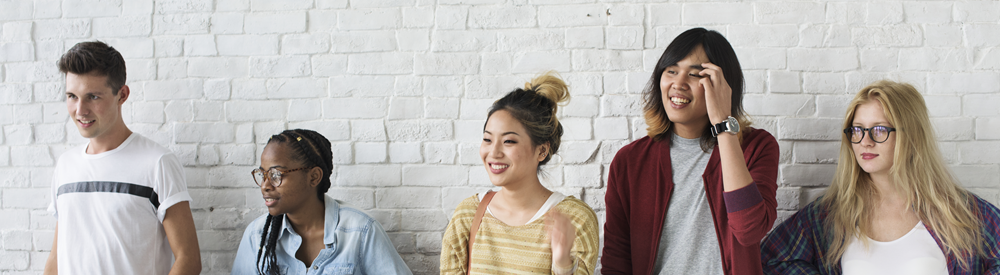 Five young adults stand casually against a white brick wall, dressed in casual, modern clothing with varied styles and colours.