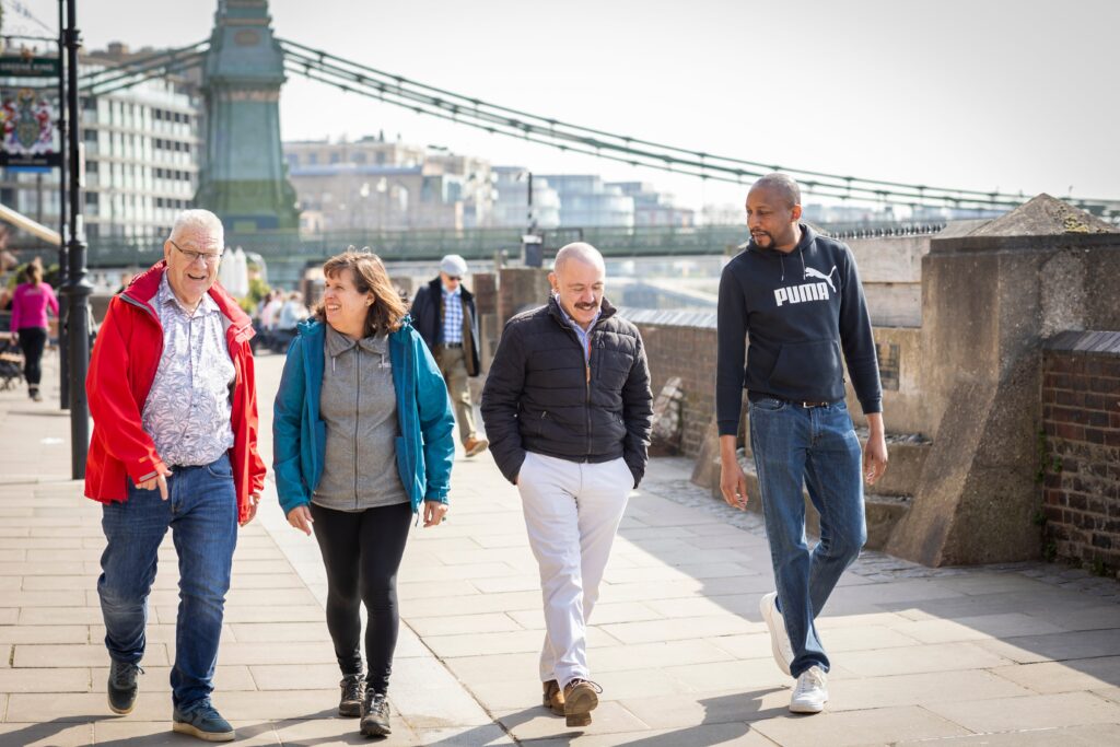 Four adults walking along a sunny riverside promenade with a suspension bridge in the background.