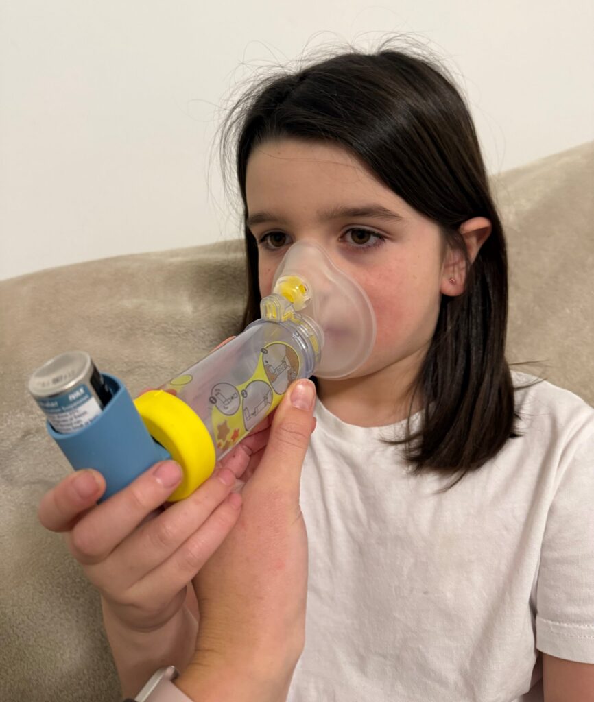 Photo of child (Ivy) being helped to use an aerochamber spacer