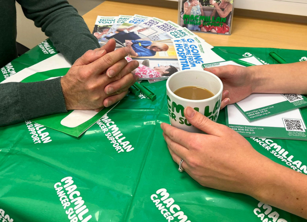 Two people sitting at a table covered with a green Macmillan Cancer Support cloth, one holding a coffee cup, the other with hands clasped.