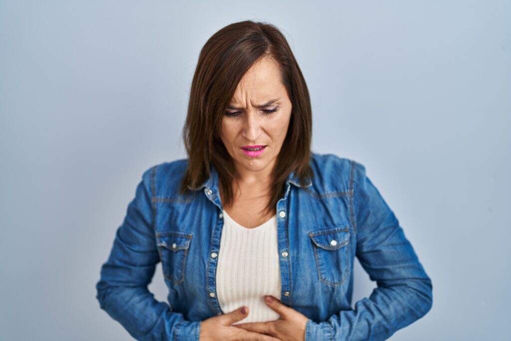 Middle age brunette woman wearing casual denim jacket over isolated background with hand on stomach because indigestion, painful illness feeling unwell. ache concept.