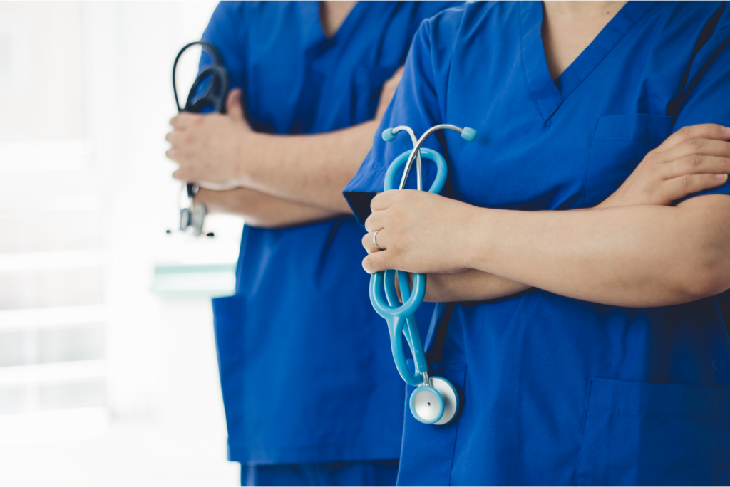 Two healthcare professionals in blue scrubs stand side by side with their arms crossed, each holding a stethoscope; their faces are not visible, focusing attention on their uniforms and medical equipment.