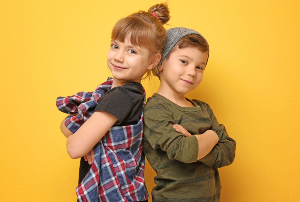 Two children stand back to back, with arms crossed. One child has a red, white, and blue plaid shirt over a black shirt, and the other wears a green striped long-sleeved shirt and a grey beanie.