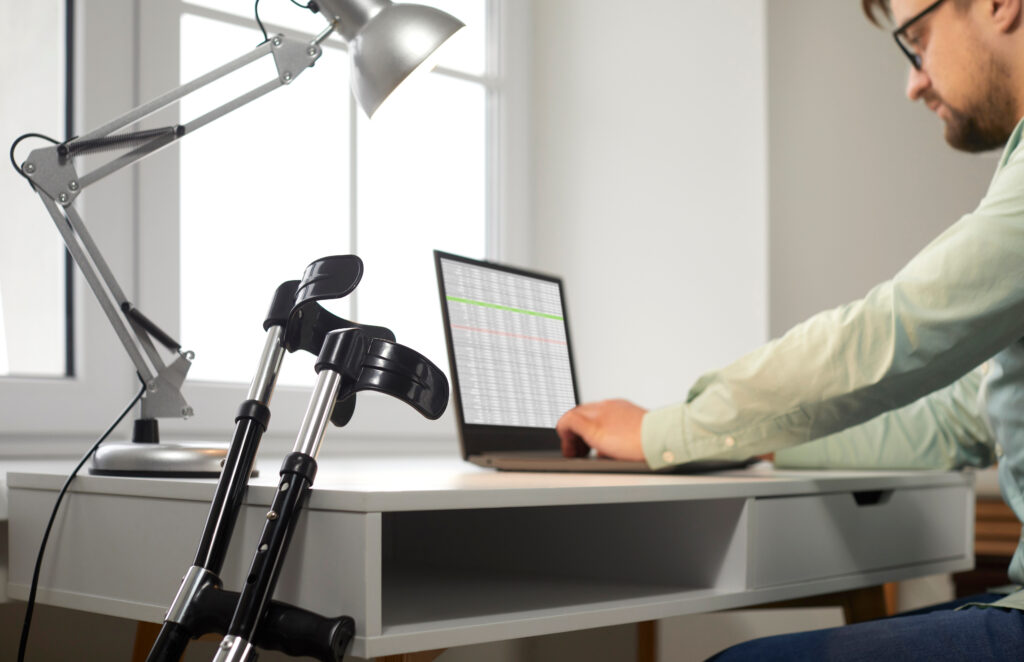 Photograph of a man wearing a green shirt sat at a desk using a laptop. A pair of crutches lean on the desk.