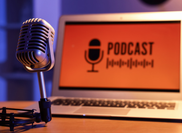 Photograph of a vintage microphone on a table, behind it is a laptop with the word 'podcast' on the screen