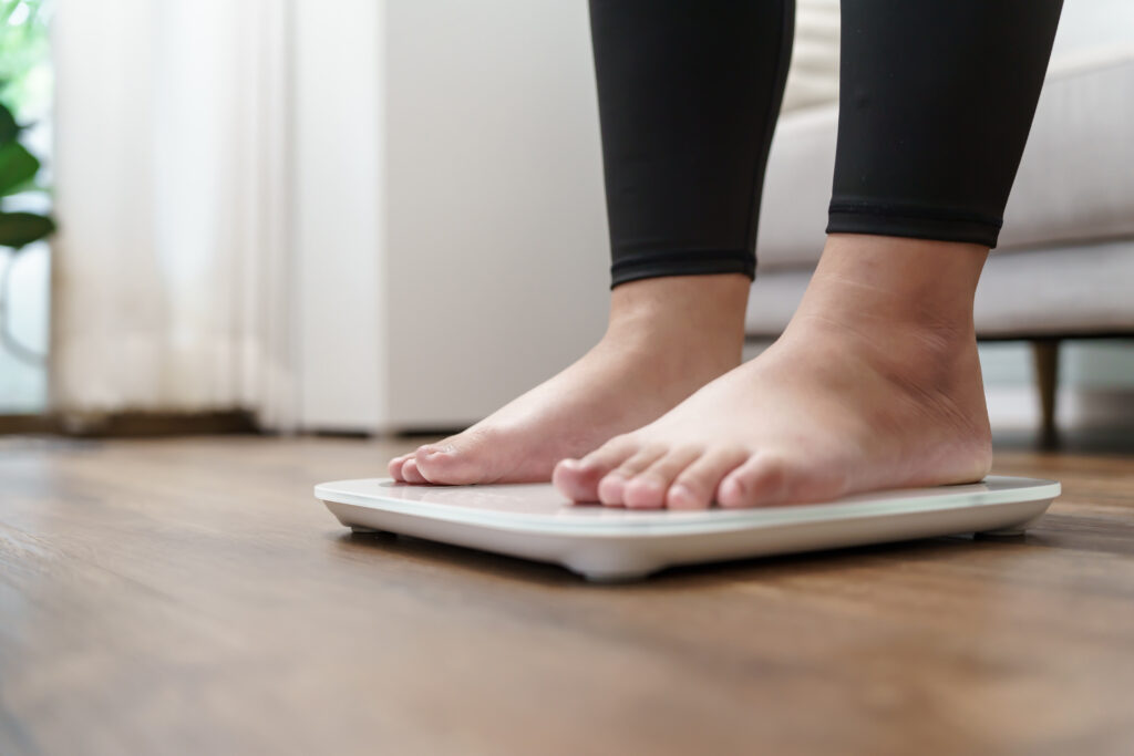 Close-up of bare feet standing on a digital weighing scale on a wooden floor in a bright indoor room. The person is wearing black leggings, with the background softly blurred.